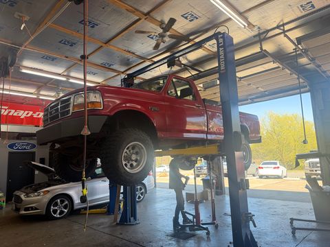 Red 1990s Ford F-250 raised on a two-post lift, with a silver Ford sedan in the next bay