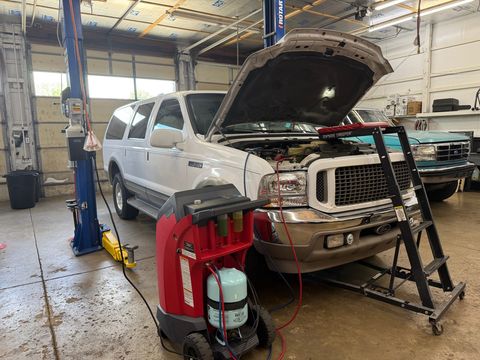White Ford Excursion with the hood open, hooked up to an A/C machine, with a teal Ford pickup behind it
